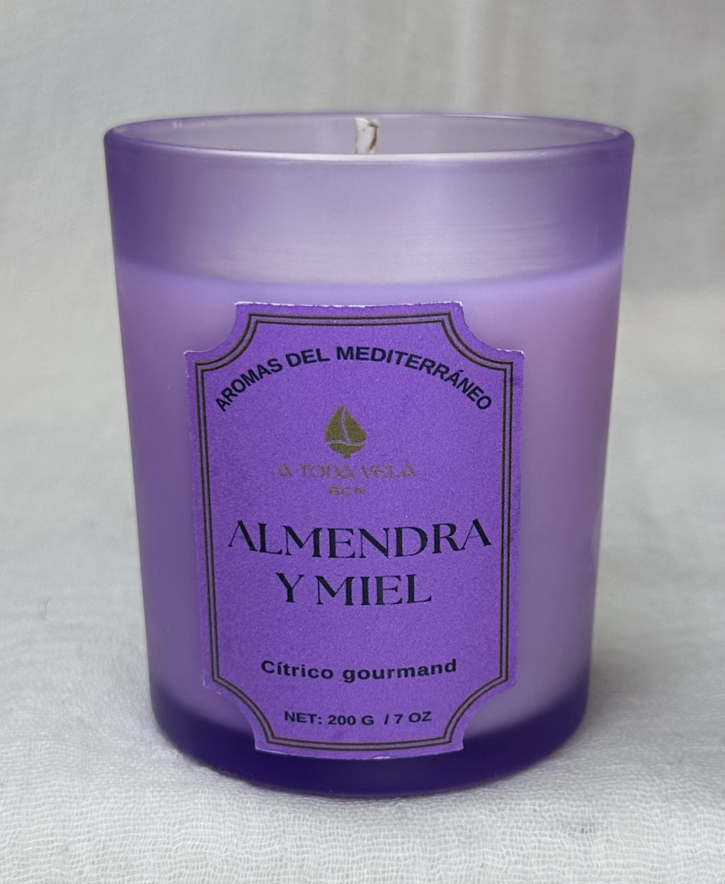 Primer plano de la vela de cera vegetal Almendra y Miel de 200g. El vaso esmerilado de color violeta claro lleva la etiqueta "Cítrico gourmand" y "AROMAS DEL MEDITERRÁNEO". Detalle de la cera de soja pura y la mecha de algodón para una quema limpia. Aroma cálido y acogedor con notas de Mandarina, Almendra y Vainilla.
