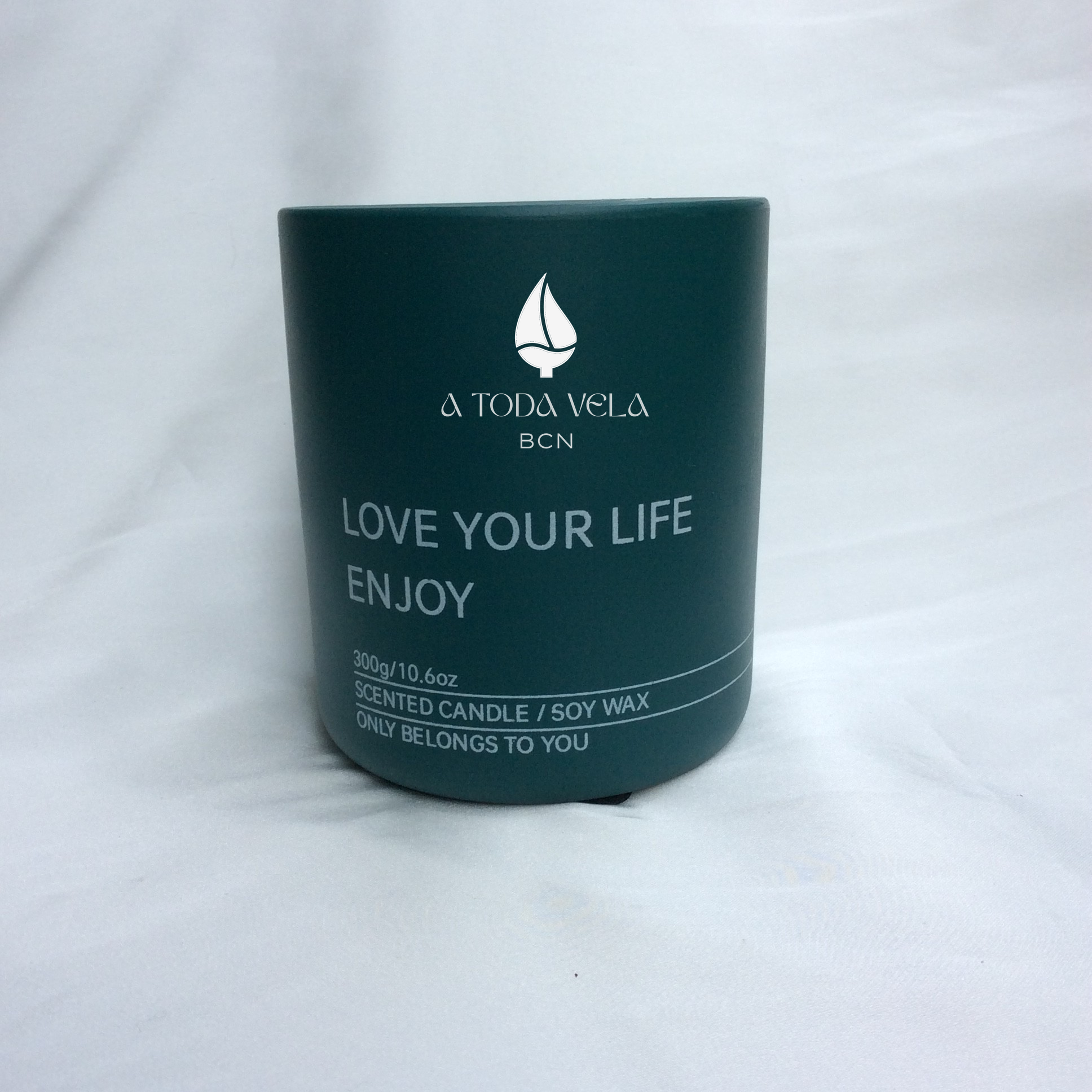 Primer plano de la vela de cera de soja 100% ecológica A Toda Vela BCN, colección Rumbo al Deseo, en recipiente color verde oscuro. Se aprecia el logo de A TODA VELA BCN y el texto "LOVE YOUR LIFE ENJOY" y la información "300g/10.6oz SCENTED CANDLE / SOY WAX". El envase elegante resalta la fragancia Oriental-Floral de Rosa Damasco y Vainilla Bourbon.