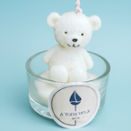 Vela blanca en forma de osito de peluche sentado dentro de un vaso de cristal, con la etiqueta de la marca "A Toda Vela BCN" en la parte frontal, sobre un fondo azul claro.