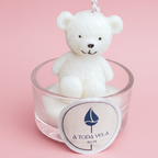 Vela blanca en forma de osito de peluche sentado dentro de un vaso de cristal, con la etiqueta de la marca "A Toda Vela BCN" en la parte frontal, sobre un fondorosadoclaro.