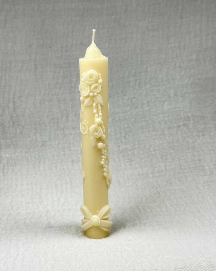 : Cirio de cumpleaños de color amarillo pálido con intrincados relieves decorativos de rosas, hojas y perlas a lo largo del cuerpo, y un lazo en la base, sobre un fondo beige claro y texturizado.
