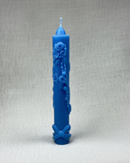 : Cirio de cumpleaños de color azul pálido con intrincados relieves decorativos de rosas, hojas y perlas a lo largo del cuerpo, y un lazo en la base, sobre un fondo beige claro y texturizado.