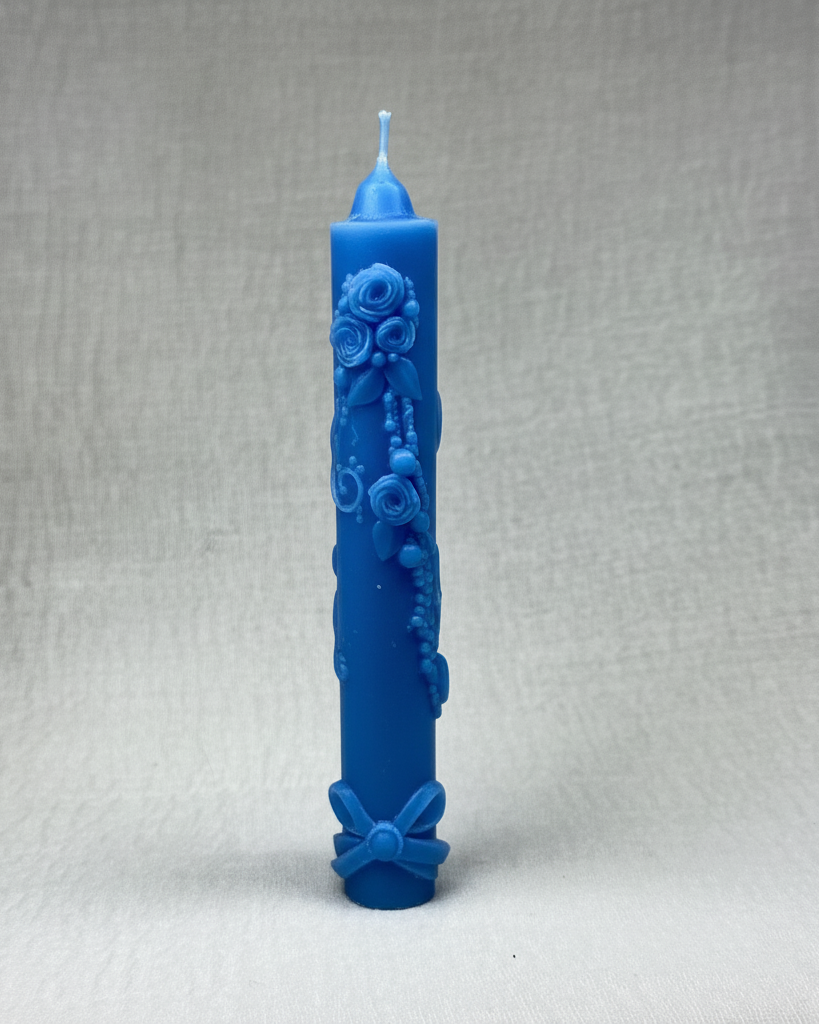 : Cirio de cumpleaños de color azul pálido con intrincados relieves decorativos de rosas, hojas y perlas a lo largo del cuerpo, y un lazo en la base, sobre un fondo beige claro y texturizado.