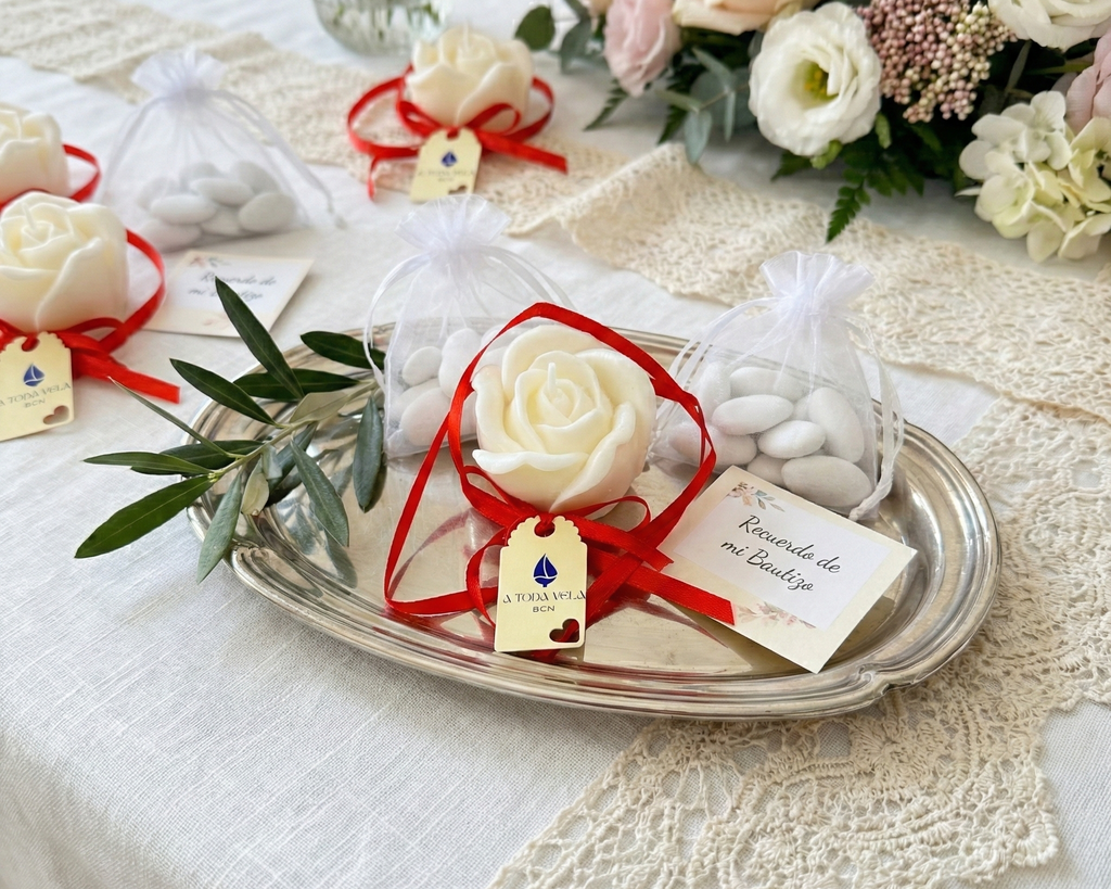 Presentación de detalles de bautizo elegantes en bandeja de plata. Incluye la vela de rosa blanca con lazo rojo de A Toda Vela BCN, bolsitas de organza con peladillas y tarjeta 'Recuerdo de mi Bautizo' sobre una mesa decorada con flores.