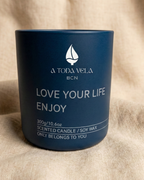 Vela aromática de soja en recipiente azul oscuro mate de la marca 'A Toda Vela BCN'. Muestra el texto blanco 'LOVE YOUR LIFE ENJOY' y está colocada sobre un fondo de tela de lino beige