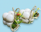 Dos velas blancas con forma de burbujas, envueltas individualmente con cuerda rústica y adornadas con pequeñas flores artificiales de colores y hojas verdes, sobre un fondo azul claro.