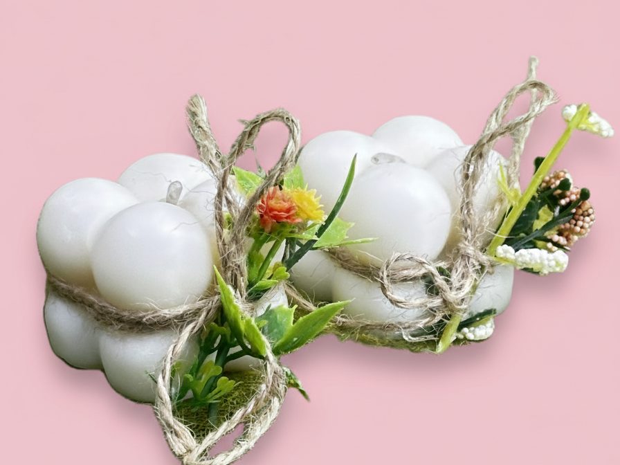 Dos velas blancas con forma de burbujas, envueltas individualmente con cuerda rústica y adornadas con pequeñas flores artificiales de colores y hojas verdes, sobre un fondo rosaclaro.