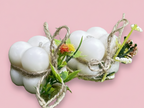 Dos velas blancas con forma de burbujas, envueltas individualmente con cuerda rústica y adornadas con pequeñas flores artificiales de colores y hojas verdes, sobre un fondo rosaclaro.