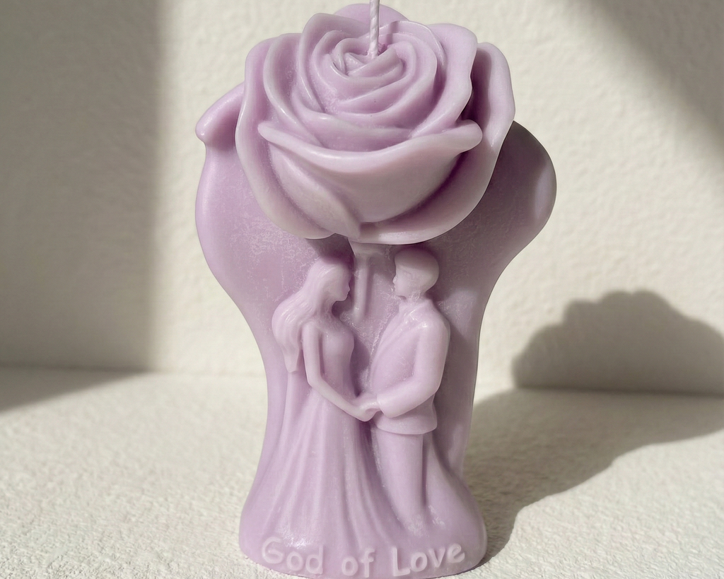 Vela recuerdo de boda con figura de pareja de novios bajo una gran rosa. Escultura de cera en color lila suave con la inscripción 'God of Love' en la base