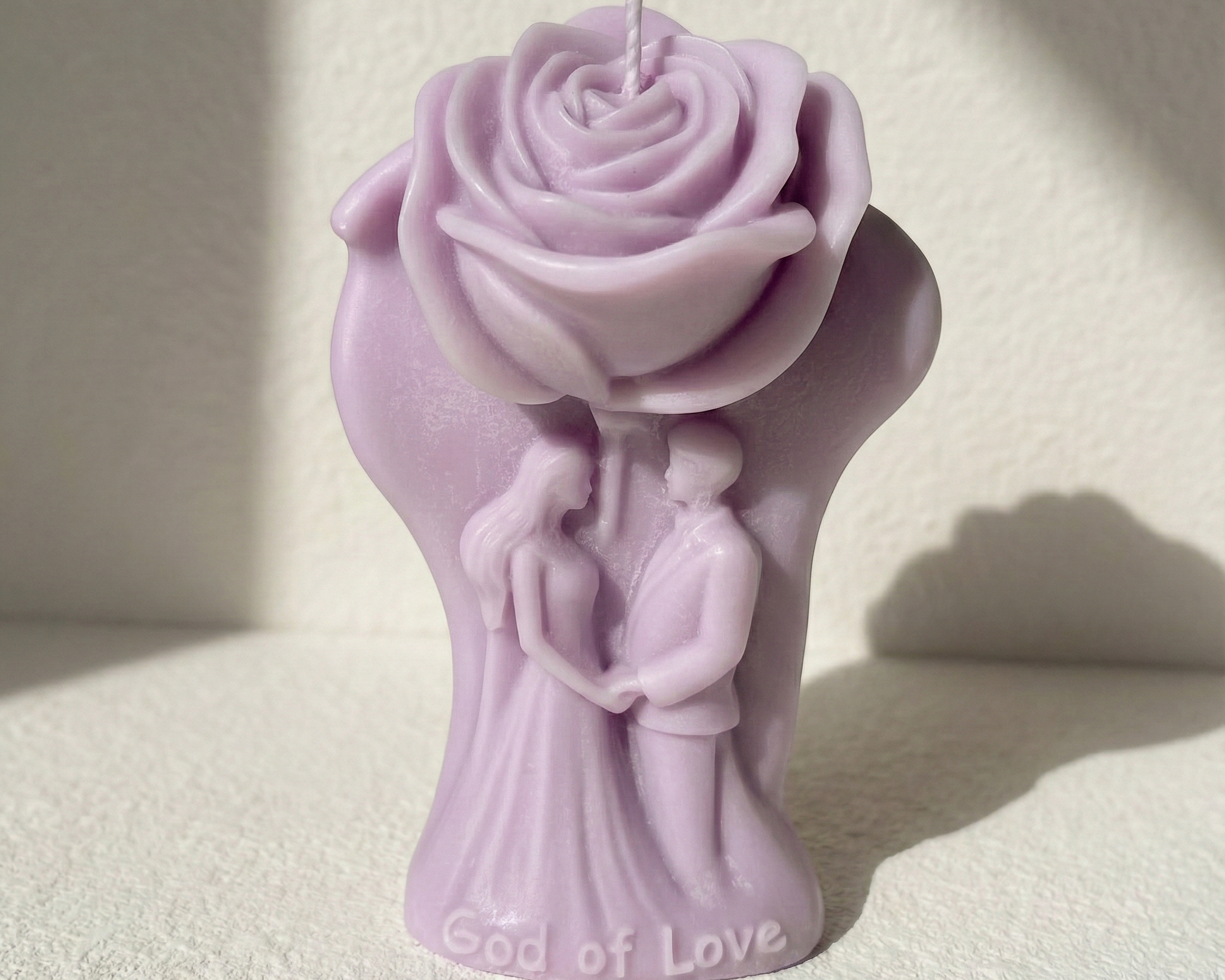 Vela recuerdo de boda con figura de pareja de novios bajo una gran rosa. Escultura de cera en color lila suave con la inscripción 'God of Love' en la base