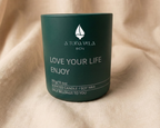Vela aromática de soja en tarro de cerámica verde mate con el logotipo de A TODA VELA BCN. La vela presenta el mensaje inspirador 'LOVE YOUR LIFE ENJOY' en letras blancas y los detalles '300g/10.6oz SCENTED CANDLE / SOY WAX ONLY BELONGS TO YOU'. La vela está colocada sobre un fondo de tela de lino beige claro con pliegues suaves.