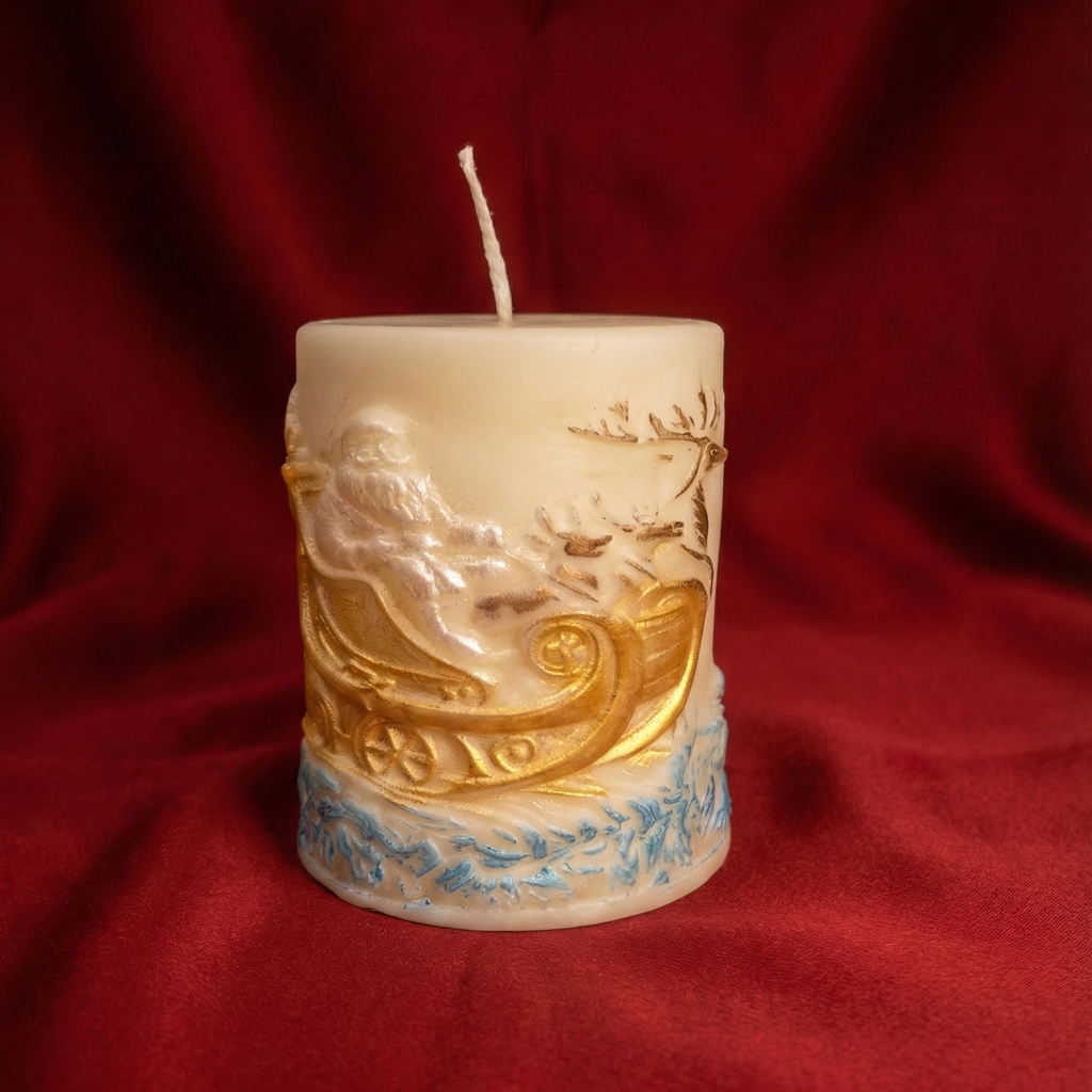 Primer plano de una vela pilar navideña de cera color marfil, decorada con un relieve detallado de Papá Noel en su trineo dorado tirado por renos y detalles azules en la base, sobre un fondo de tela roja suave.