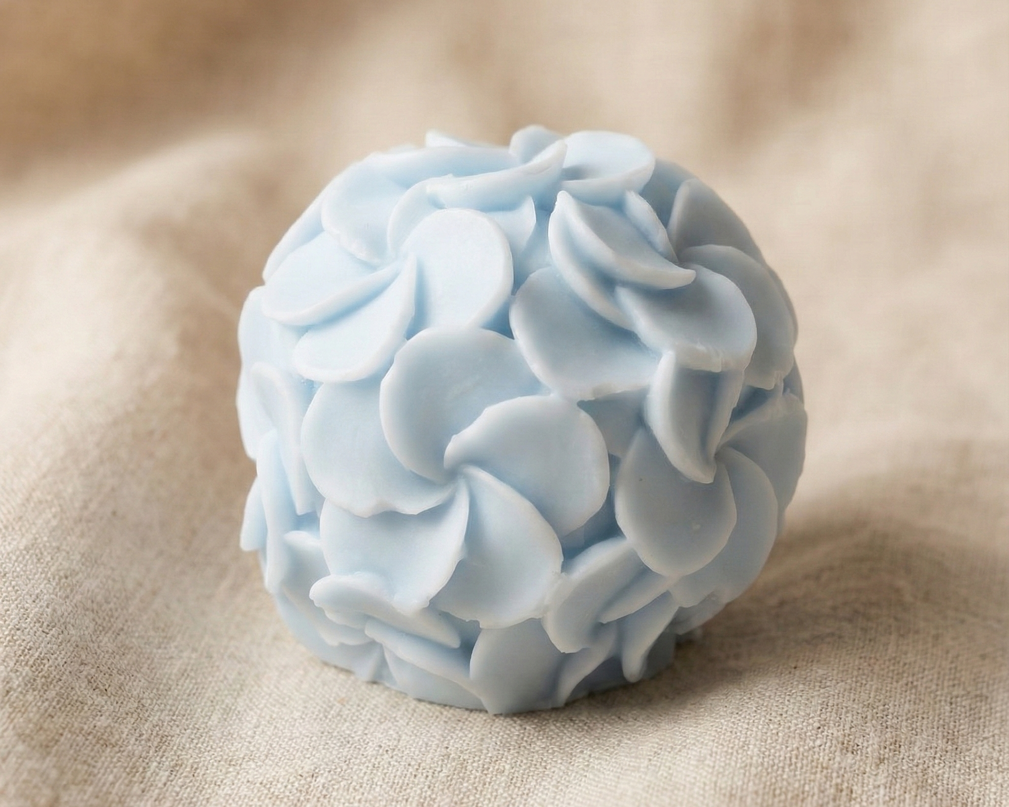 Primer plano de una vela redonda artesanal en tono azul pastel, cubierta completamente por un diseño detallado de pequeñas flores de cera sobre un fondo de lino beige.