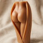 Vela decorativa escultural con forma de glúteos y piernas femeninas emergiendo de una envoltura orgánica texturizada. La vela tiene un tono de piel mestiza cálido y está situada sobre un fondo de tela beige suave.