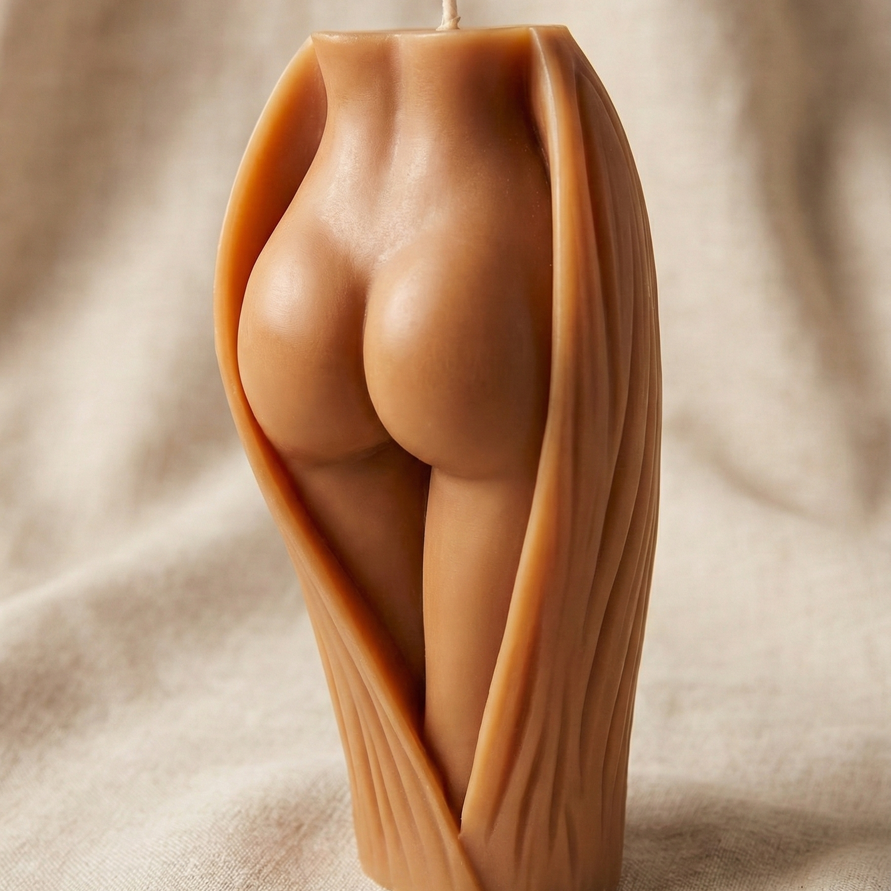 Vela decorativa escultural con forma de glúteos y piernas femeninas emergiendo de una envoltura orgánica texturizada. La vela tiene un tono de piel mestiza cálido y está situada sobre un fondo de tela beige suave.