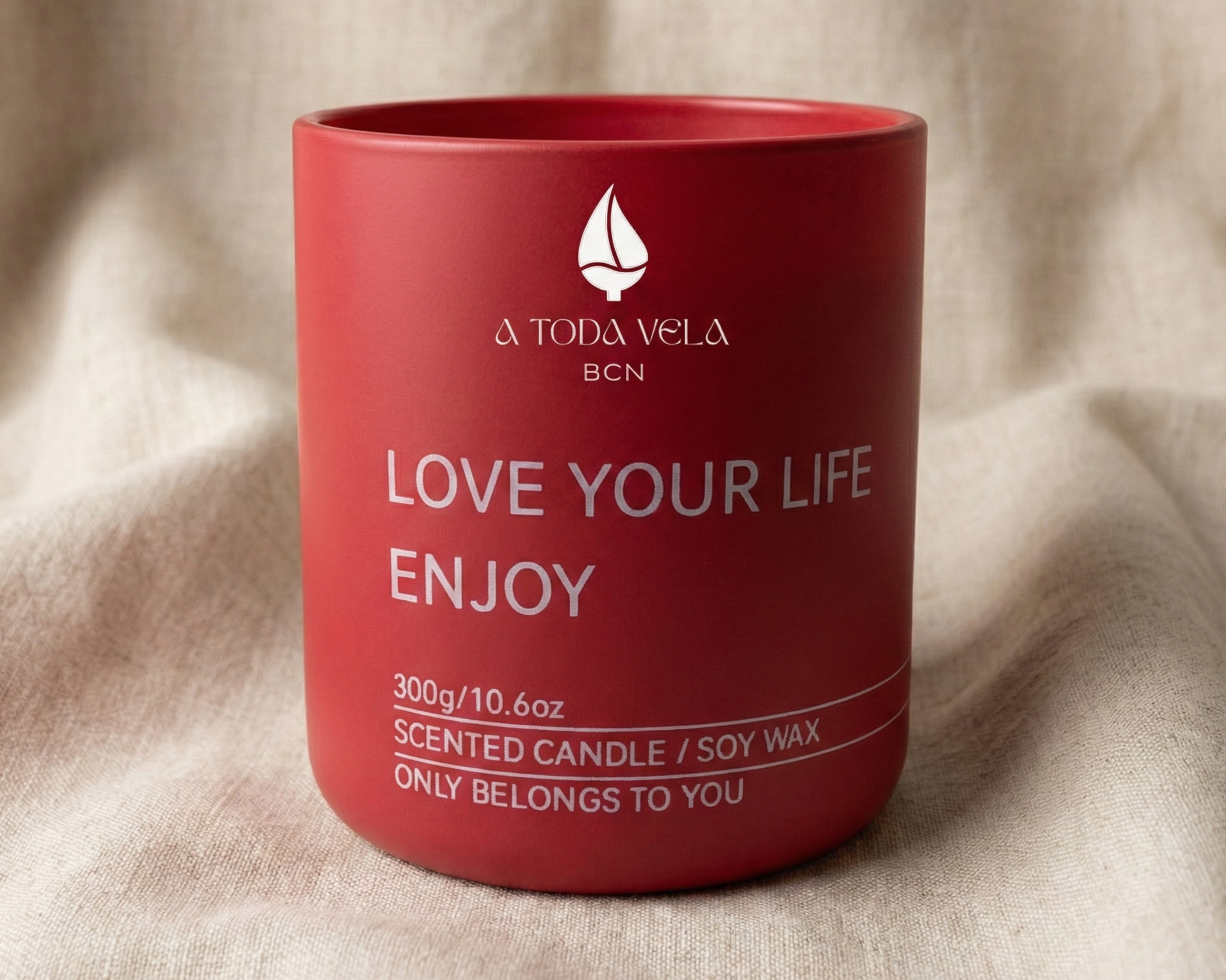 Vela aromática de soja en recipiente de color rojo mate de la marca 'A Toda Vela BCN'. Muestra el mensaje inspirador 'LOVE YOUR LIFE ENJOY' en letras blancas y detalles del peso (300g). La vela está situada sobre un fondo de tela suave color beige claro."