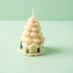 Fotografía de producto en primer plano de una vela decorativa navideña. La vela tiene forma de árbol de Navidad blanco o pino nevado hecho de capas de cera color crema. En la base, presenta detalles en relieve de pequeñas casas y árboles de color verde. Está situada sobre una superficie y fondo liso de color verde menta claro.