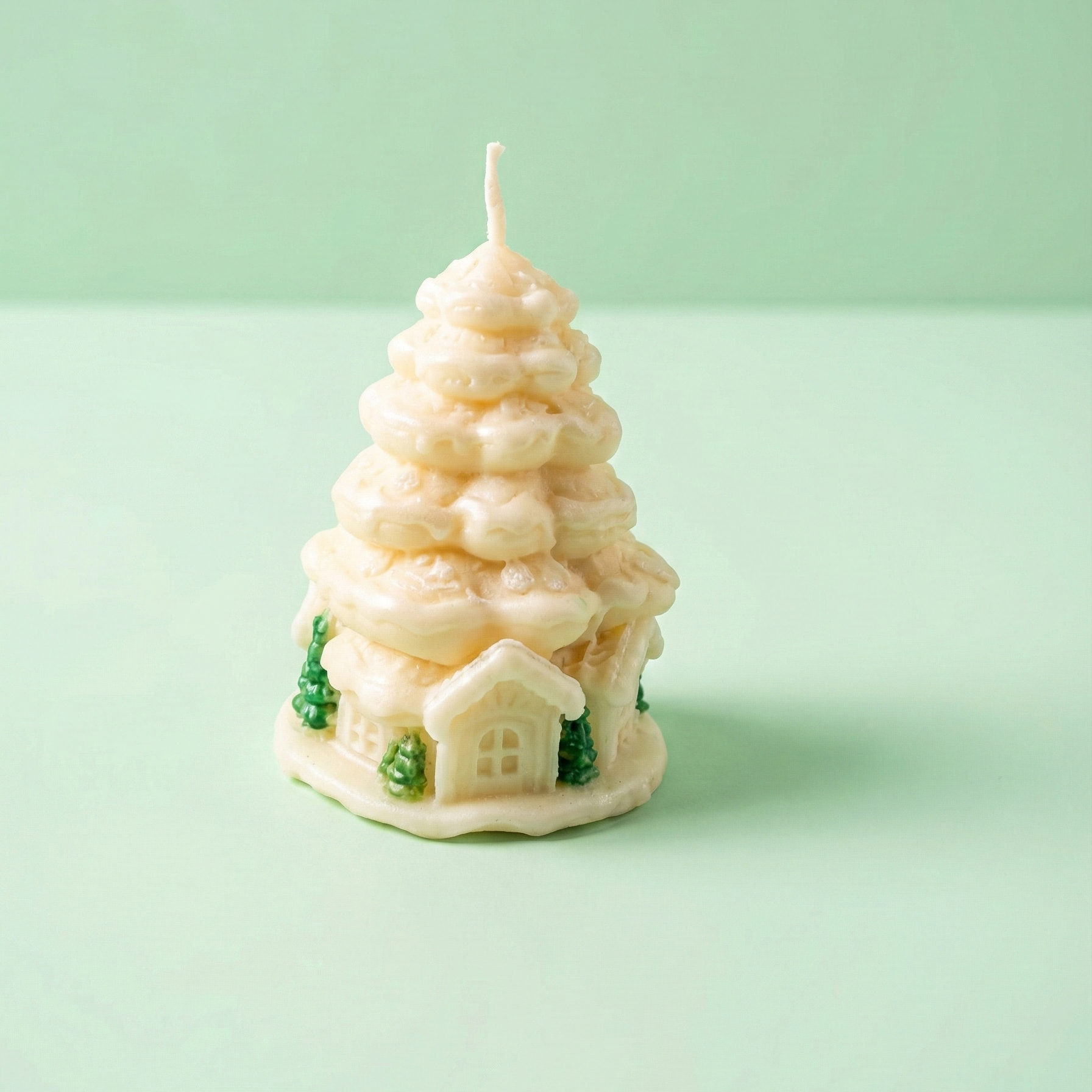 Fotografía de producto en primer plano de una vela decorativa navideña. La vela tiene forma de árbol de Navidad blanco o pino nevado hecho de capas de cera color crema. En la base, presenta detalles en relieve de pequeñas casas y árboles de color verde. Está situada sobre una superficie y fondo liso de color verde menta claro.