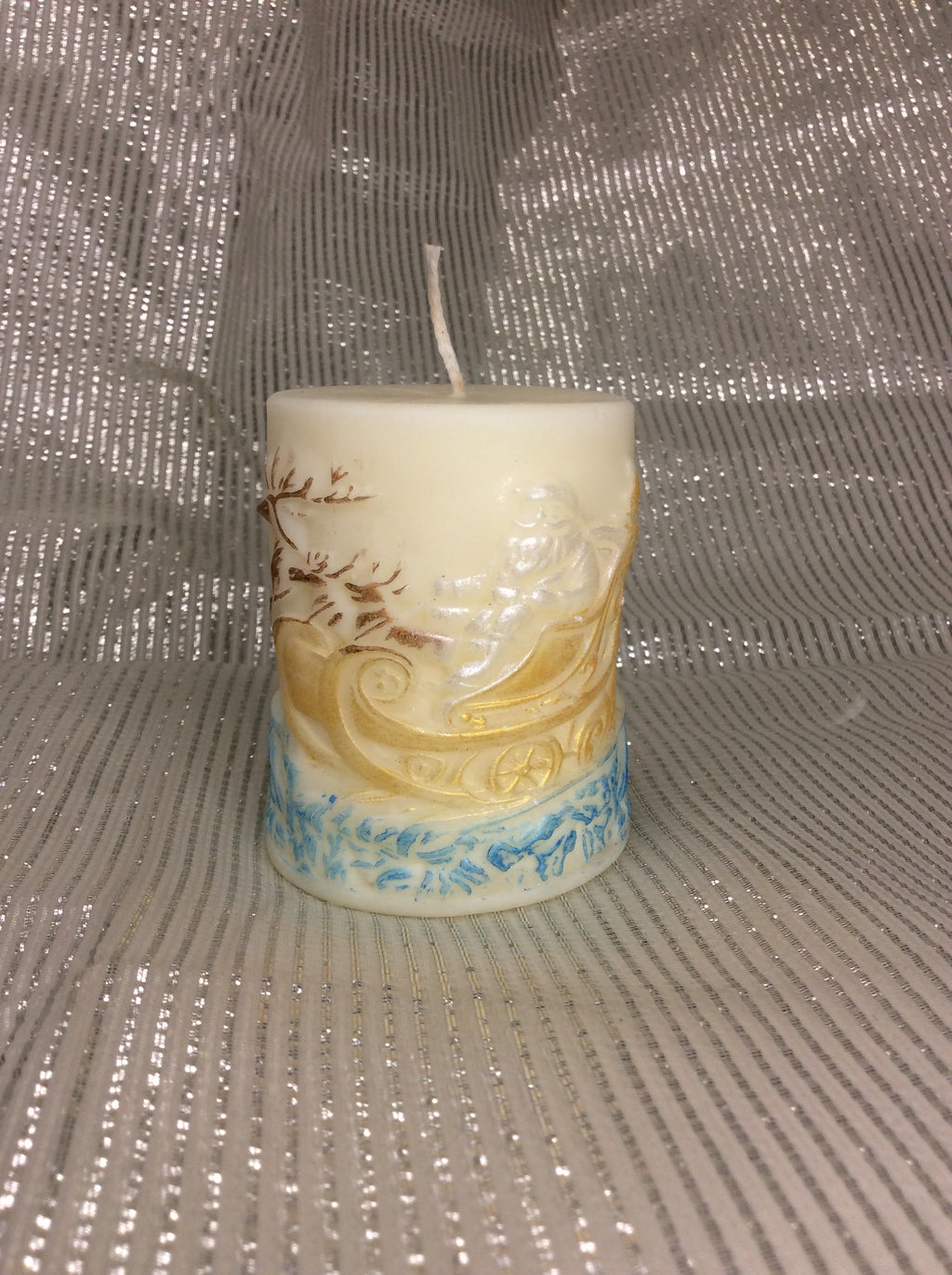 Vela Papá Noel Trineo, cilíndrica, relieve 3D pintado a mano en tonos dorados y azules sobre fondo plateado.