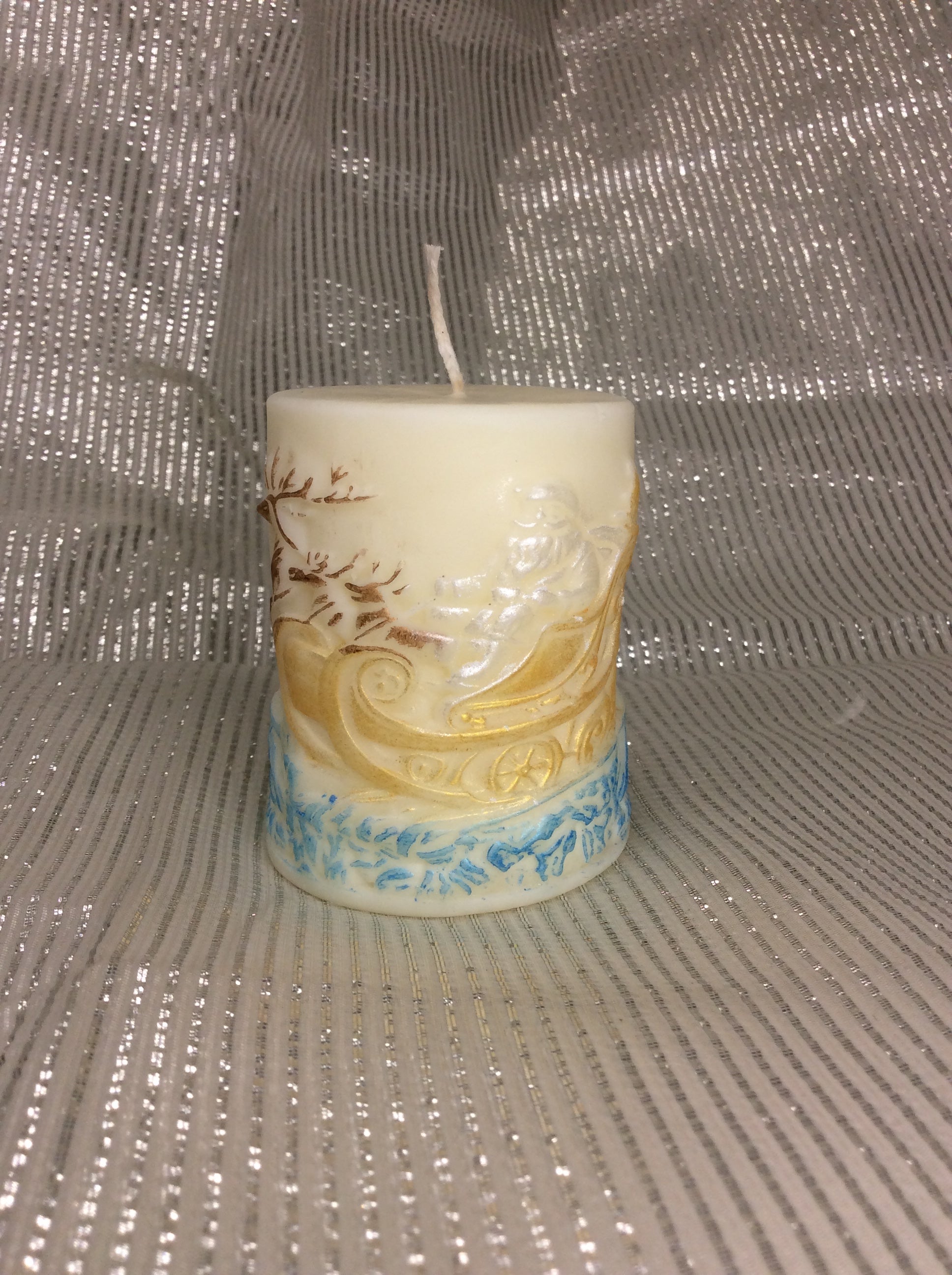 Vela Papá Noel Trineo, cilíndrica, relieve 3D pintado a mano en tonos dorados y azules sobre fondo plateado.