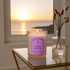 Vela perfumada de lujo Citrus Soleil de A Toda Vela BCN, encendida sobre una mesa de cristal en un primer plano. El envase de cristal esmerilado violeta claro destaca el aroma Cítrico y Floral aterciopelado de Limón Siciliano, Azahar y Tomillo. El fondo muestra un atardecer sobre el mar Mediterráneo, evocando un ambiente fresco, limpio y estimulante. Producto de cera vegetal ecológica hecho en Barcelona