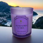 Vela aromática de lujo Almendra y Miel de A Toda Vela BCN en vaso violeta claro sobre una terraza con vistas a la costa del Mediterráneo al atardecer. La fragancia Cítrico Gourmand combina Almendra cremosa y Miel para un ambiente cálido, acogedor y pet friendly. Producto de cera vegetal ecológica de Barcelona.
