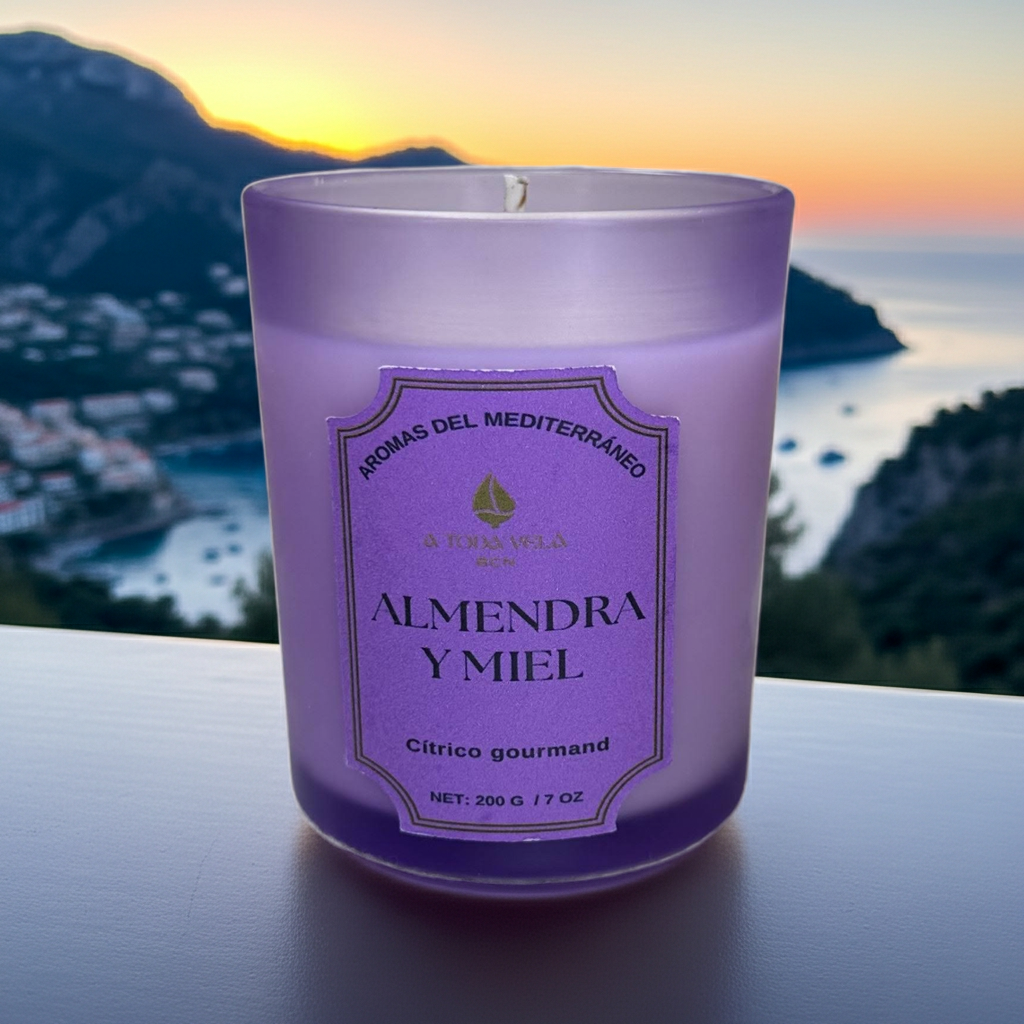 Vela aromática de lujo Almendra y Miel de A Toda Vela BCN en vaso violeta claro sobre una terraza con vistas a la costa del Mediterráneo al atardecer. La fragancia Cítrico Gourmand combina Almendra cremosa y Miel para un ambiente cálido, acogedor y pet friendly. Producto de cera vegetal ecológica de Barcelona.