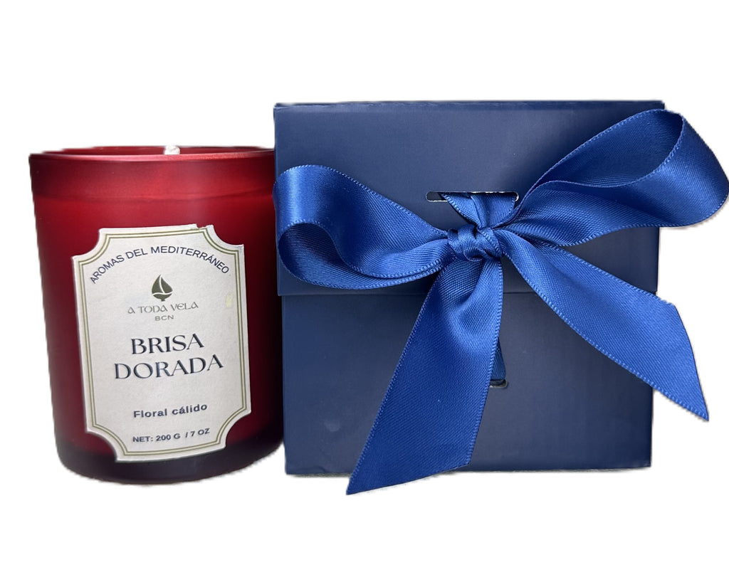 Set de regalo de la vela Brisa Dorada de A Toda Vela BCN. La vela de 200g en vaso rojo está junto a una caja de regalo azul marino atada con un lazo de raso. Ideal para bodas, bautizos, hoteles y spas, este detalle de lujo se puede personalizar y contiene un aroma Marino Floral con cera vegetal pura y pet friendly.