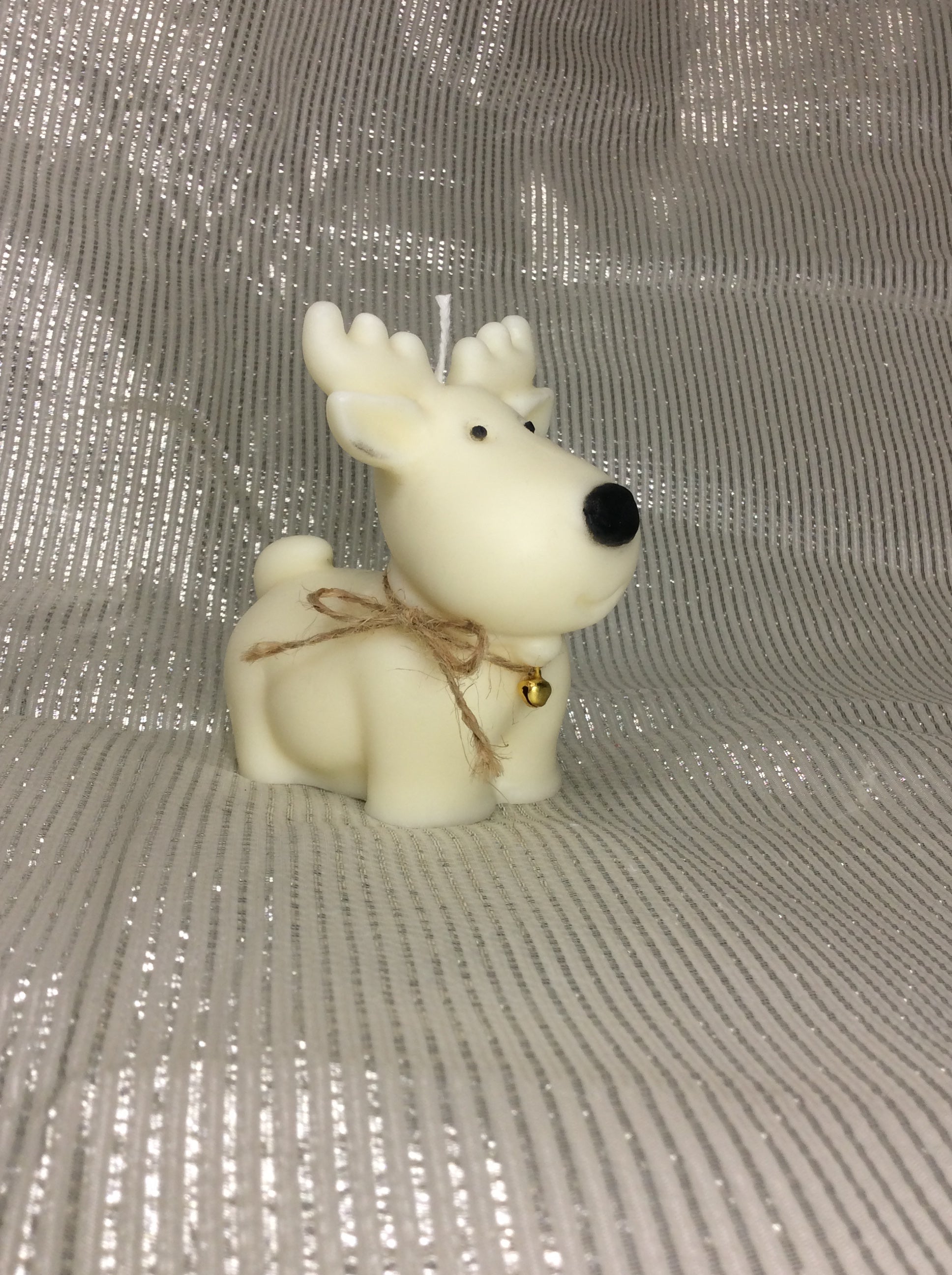 Vela Reno de Navidad con nariz negra, campana de cascabel y lazo de yute, sobre fondo plateado.