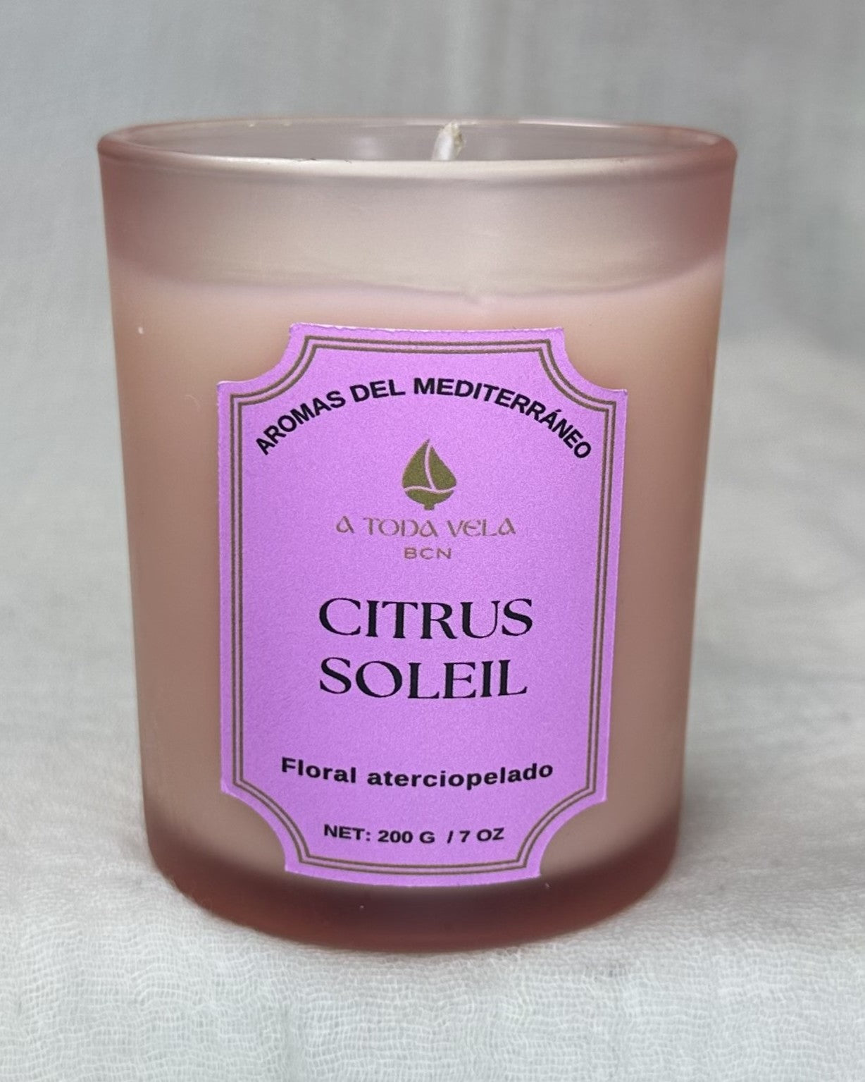 Primer plano del vaso de la vela Citrus Soleil de A Toda Vela BCN, de cera vegetal ecológica en un envase de cristal esmerilado rosa claro/pálido. La etiqueta violeta detalla el nombre del producto, "AROMAS DEL MEDITERRÁNEO" y el perfil "Floral aterciopelado". El vaso de 200g/7oz presenta la fragancia fresca de Limón Siciliano, Azahar y Tomillo.