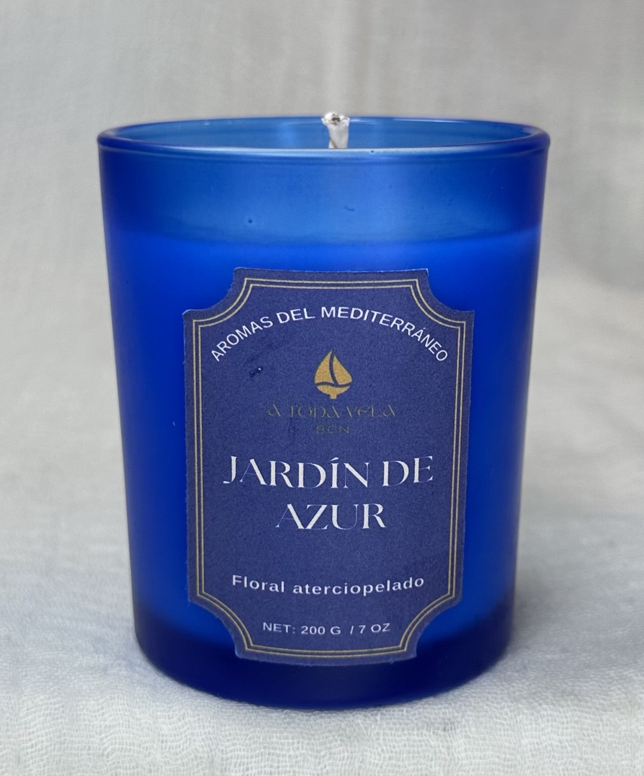 Primer plano de la vela de cera de soja Jardín de Azur de 200g en un vibrante vaso de cristal azul profundo. La etiqueta detalla "AROMAS DEL MEDITERRÁNEO" y el perfil "Floral aterciopelado". El aroma, hecho a mano en Barcelona, tiene notas iniciales de Neroli y Naranja Amarga y un fondo de Vetiver.