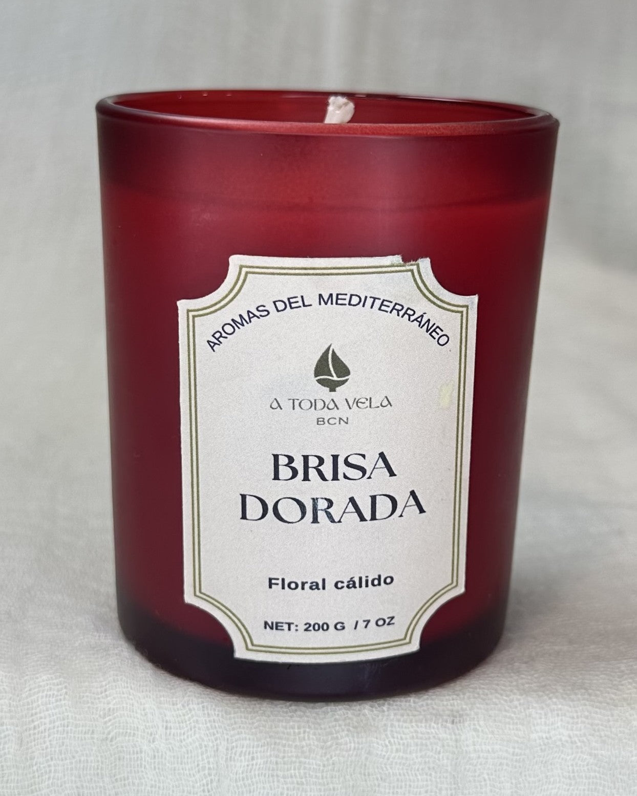 Primer plano de la vela de cera vegetal Brisa Dorada de 200g en un vaso de cristal rojo mate. La etiqueta vintage muestra "AROMAS DEL MEDITERRÁNEO" y el perfil "Floral cálido". Esta vela 100% ecológica hecha a mano en Barcelona difunde un aroma sofisticado de Flor de Naranjo, Neroli y un fondo de Cedro.