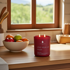 Vela perfumada de lujo A Toda Vela BCN Rumbo al Alba, encendida sobre una encimera de madera en una cocina luminosa, junto a un bol de frutas. El color rojo vibrante del envase combina con la fragancia Cítrica de Bergamota y Azahar que evoca un nuevo comienzo y frescura limpia. La escena transmite calma y serenidad a la luz de la mañana.