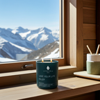 Vela perfumada de lujo A Toda Vela BCN Rumbo al Deseo en tarro verde oscuro con el mensaje "LOVE YOUR LIFE ENJOY" encendida en una mesa de madera. Fondo borroso de un balcón con barandilla y montañas nevadas al atardecer. La escena evoca un ambiente sensual y de confort.