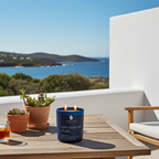 Vela perfumada de lujo A Toda Vela BCN Rumbo al Horizonte, encendida en una mesa de madera en una terraza con vistas al mar Mediterráneo y colinas. La vela de cera de soja azul marino evoca el frescor del Acorde Marino y el equilibrio de la Flor de Loto. Perfecta para un ambiente de calma y serenidad.