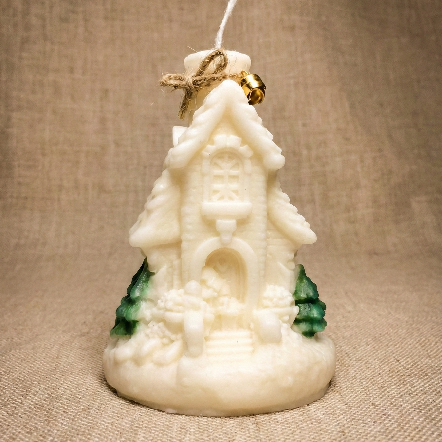 Vela decorativa navideña con forma de casita nevada, hecha de cera blanca con detalles de pinos verdes y adornada con un pequeño lazo de cuerda y una campana dorada en la parte superior.
