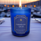 Vela de lujo Jardín de Azur de A Toda Vela BCN encendida en una mesa de restaurante, con el puerto deportivo al fondo durante el anochecer. El envase de cristal azul profundo refleja la elegancia del aroma Floral aterciopelado con notas de Neroli, Jazmín y Sándalo. Ideal para cenas o eventos sofisticados.